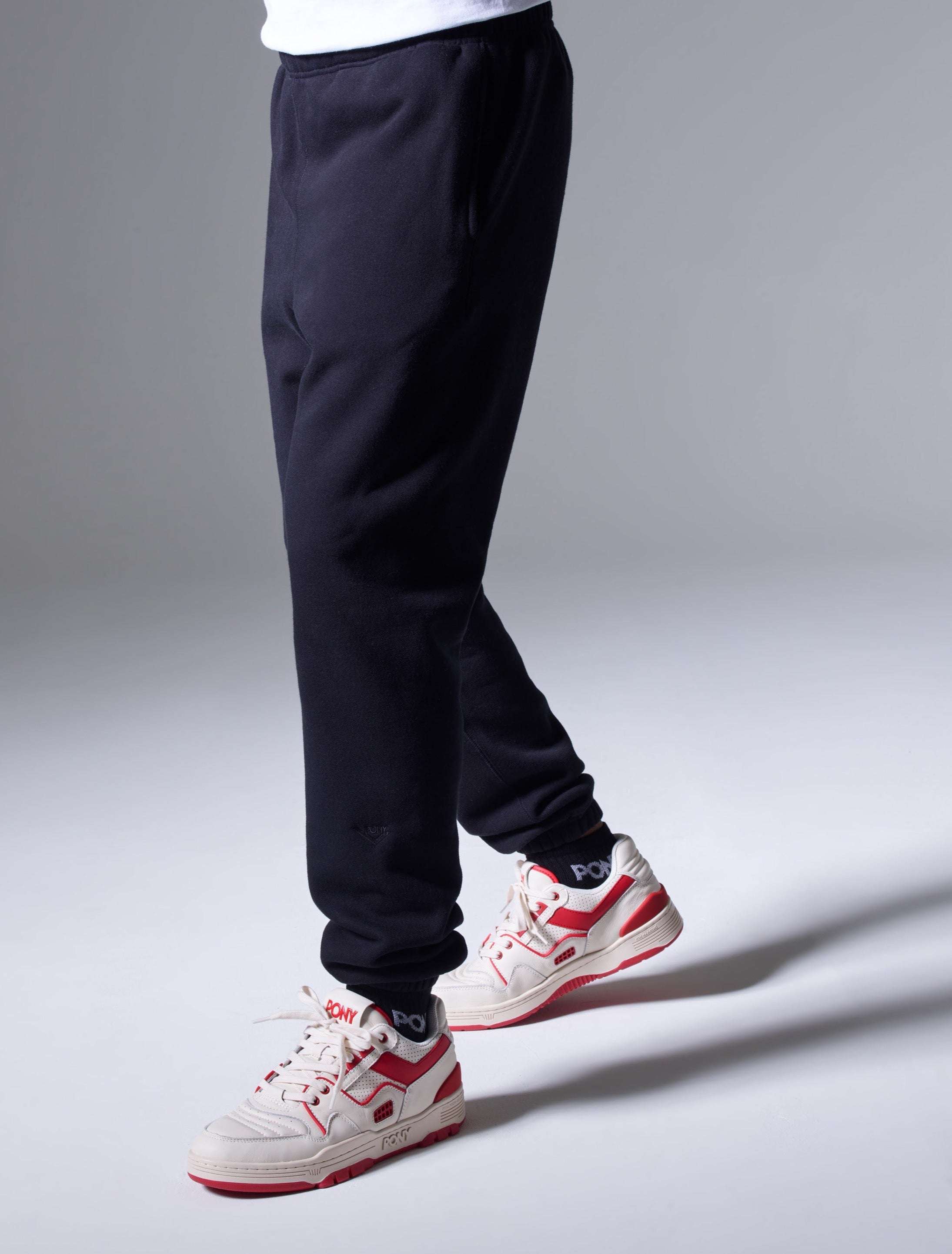 A side shot of a male model wearing PONY Jogger pants with PONY red low top M-100s.