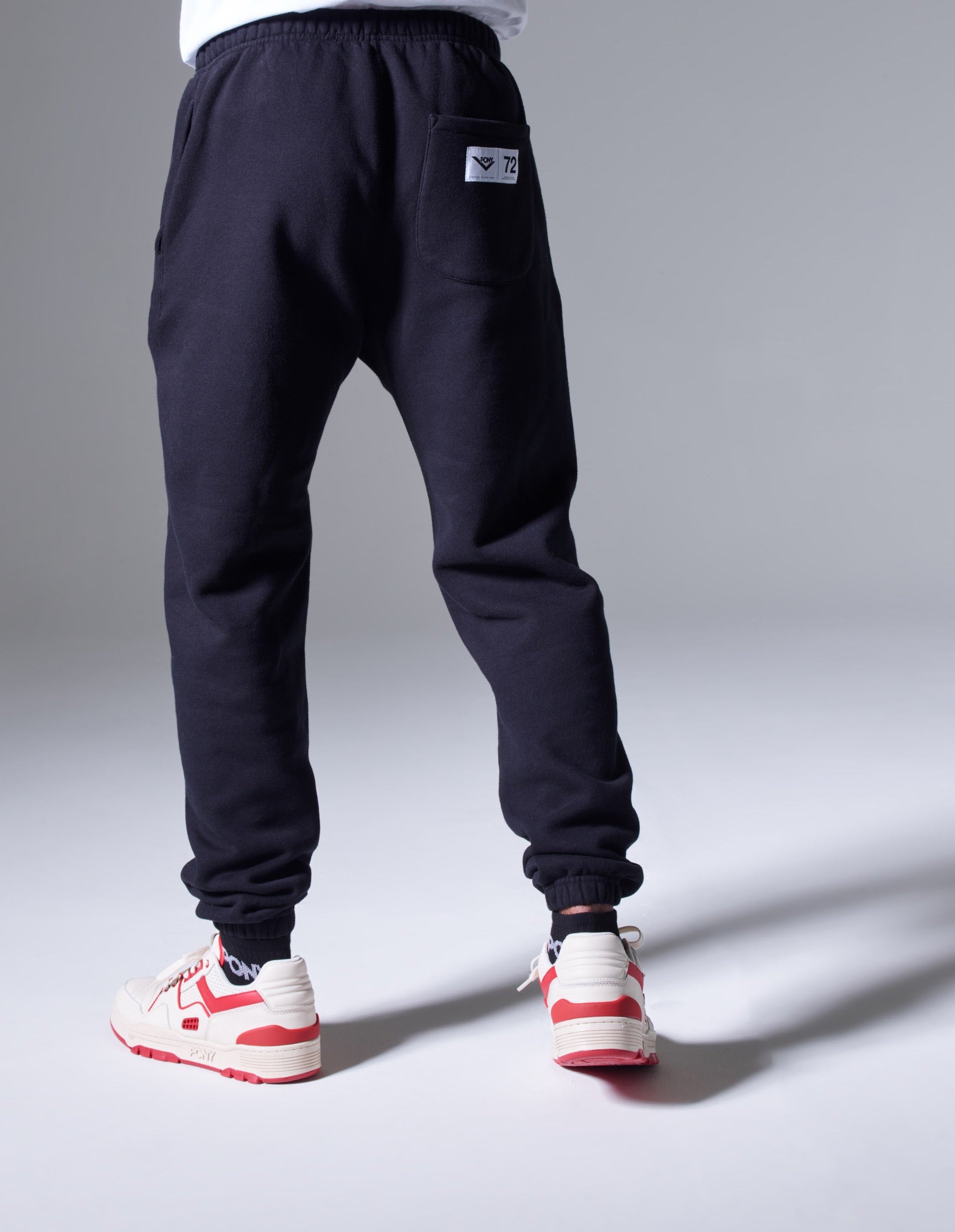 A back shot of a male model wearing PONY Jogger pants with PONY red low top M-100s. The joggers feature a right hand pocket with a PONY locker label sewn in with black "PONY" lock-up with chevron and "72" on the right.